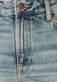 Ljusblå denimtyg med synlig indigo blekning, orange sömmar och en metallknapp vid midjan; har en design med lapplåda.