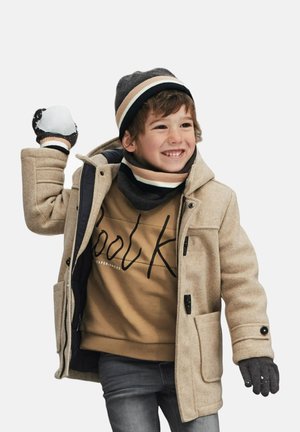 Cappotto di lana beige con due tasche frontali e bottoni neri, indossato sopra un felpa marrone. Cappello e sciarpa grigi in maglia con strisce. Guanti grigi.