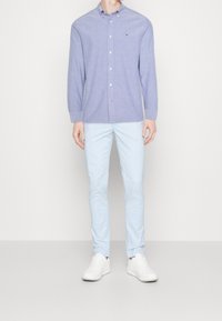 Muž stojí oblečený v světle modrých slim kalhotách, bílých teniskách a dlouhém světle fialovém košili s knoflíky a malým logem na hrudi.