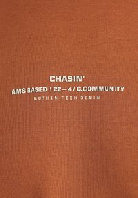 Tkanina v odstínech rzi s bílým textem. Text zahrnuje "CHASIN'," "AMS BASED / 22-4 / C.COMMUNITY," a "AUTHEN-TECH DENIM." Hladký povrch.