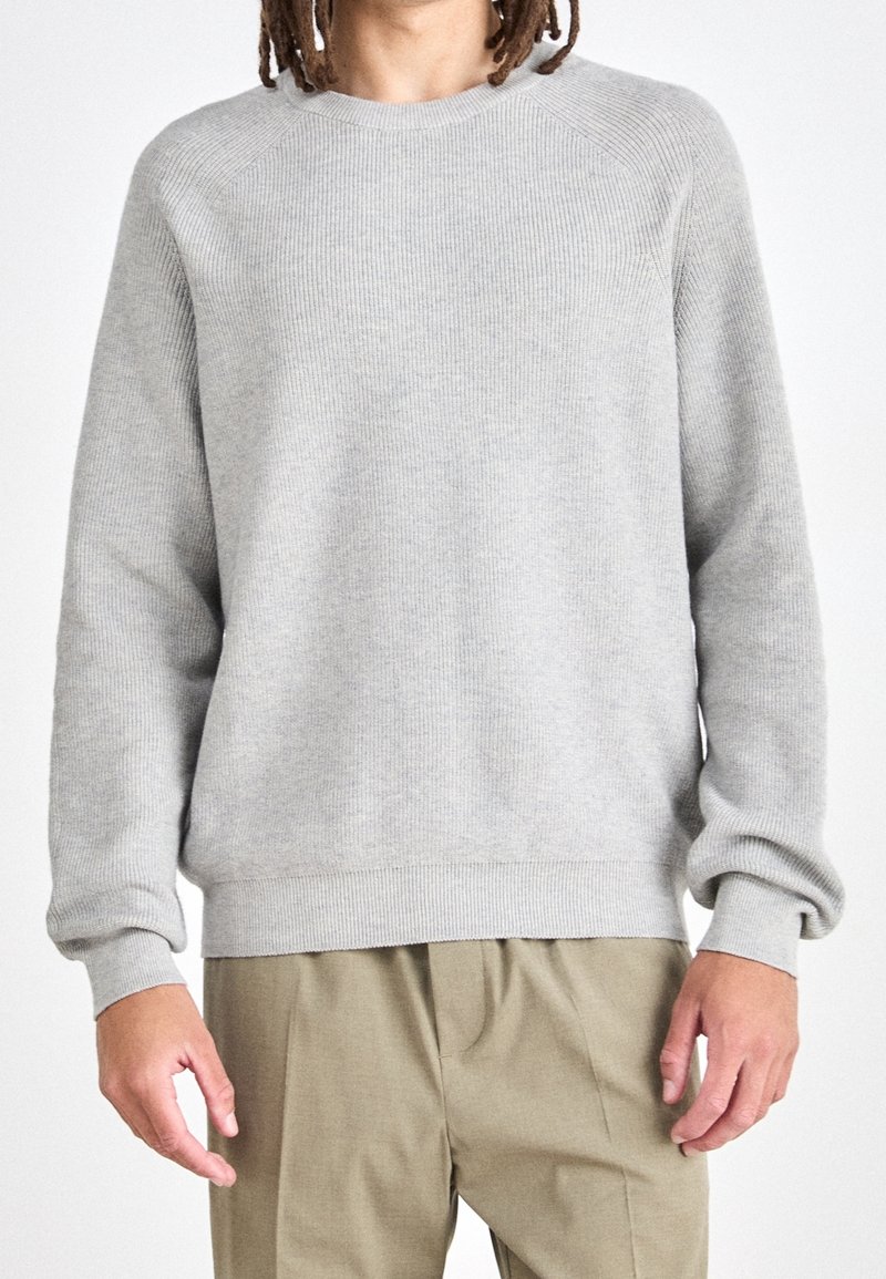 Ljusgrå crewneck-tröja med ribbat mönster, långa ärmar och avslappnad passform. Kombinerad med beige byxor. Minimalistisk design.