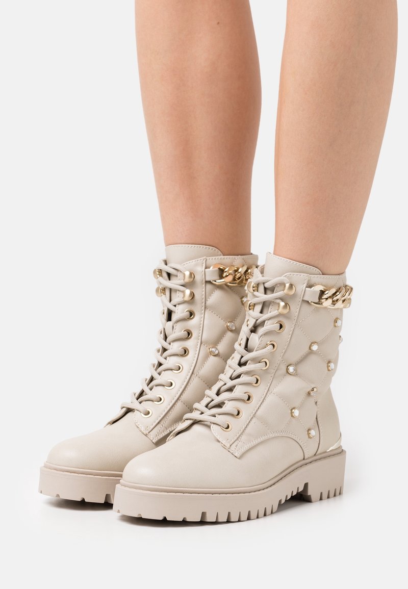 Bottines beiges à lacets avec un design matelassé, des clous dorés et un détail décoratif en chaîne dorée portées sur des jambes nues.