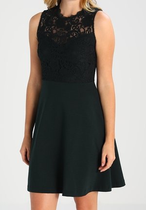 Femme portant une robe noire sans manches avec un corsage en dentelle à motif floral et une jupe unie, debout devant un fond clair.