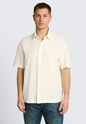 Mann trägt ein schlichtes weißes kurzärmeliges Hemd mit Knöpfen und blaue Jeans, steht vor einem hellgrauen Hintergrund.