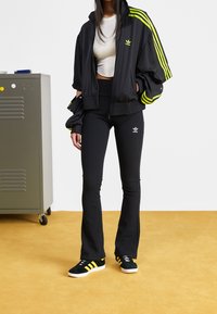 Svart sportjacka med neongula ränder, kombinerad med svarta utsvängda leggings. Svarta sneakers med gula accenter.