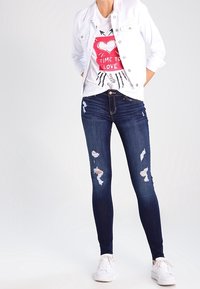 Jeans ajustados de color azul oscuro con detalles desgastados, combinados con una chaqueta de mezclilla blanca sobre una camiseta gráfica. Zapatillas blancas completan el conjunto.