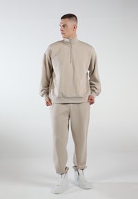 Beige zip-up sweatshirt with a high collar, loose fit, and ribbed cuffs, paired with matching tapered sweatpants and white high-top sneakers.