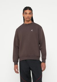 Joven con pelo trenzado que lleva sudadera Jordan marrón oscura y pantalones negros con acentos rojos, de pie contra un fondo blanco.
