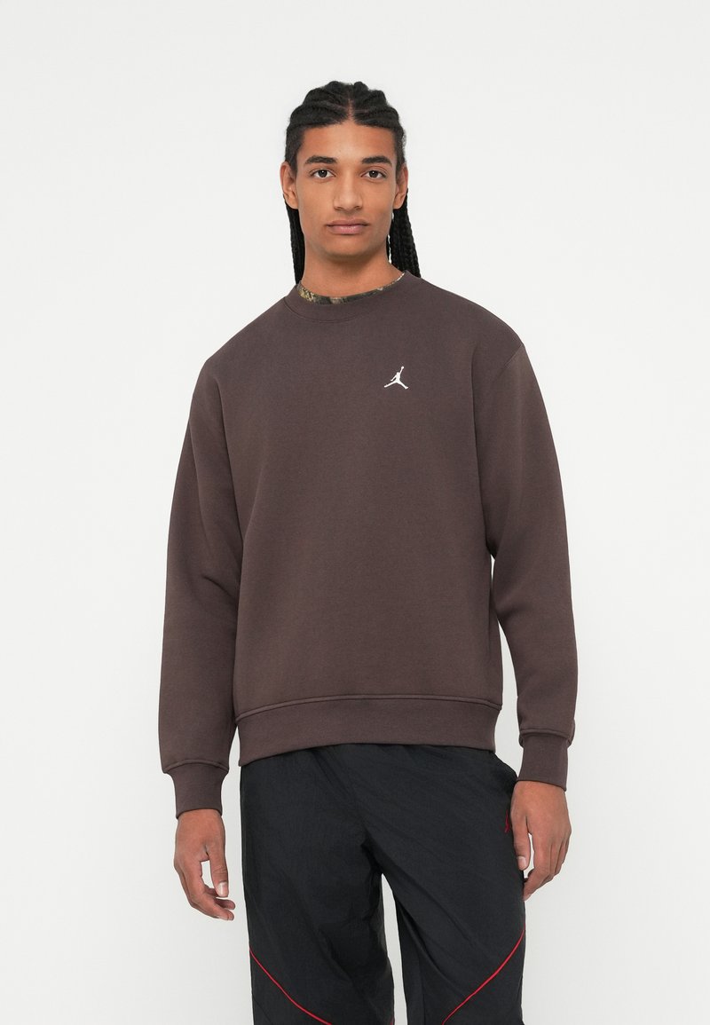 Joven con pelo trenzado que lleva sudadera Jordan marrón oscura y pantalones negros con acentos rojos, de pie contra un fondo blanco.