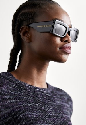 Óculos de sol oversized pretos com formato retangular, lentes reflexivas prateadas e marcação em dourado "Alexander McQueen" na armação.