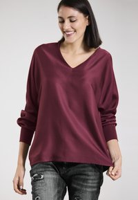 Burgundy v-neck top with long sleeves and loose fit, made of smooth material, paired with distressed gray jeans.