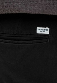 Crni platneni džep s ušivenim rubom, s bijelom etiketom na kojoj piše "JACK & JONES EST. 1990." Tekstura sive tkanine u pozadini.