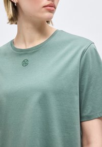 Κοντινό πλάνο ενός ατόμου που φοράει πράσινο t-shirt με στρογγυλή λαιμόκοψη και μικρό σκούρο πράσινο κυκλικό λογότυπο στο στήθος, με κοντά ξανθά μαλλιά και ασημένια σκουλαρίκια κρίκους.