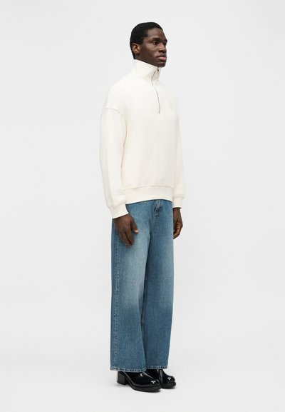 Άνδρας που στέκεται φορόντας εκρού φούτερ με φερμουάρ στο λαιμό, φαρδιά ξεβαμμένα μπλε τζιν και μαύρα παπούτσια με τακούνι, μπροστά σε απλό φόντο.