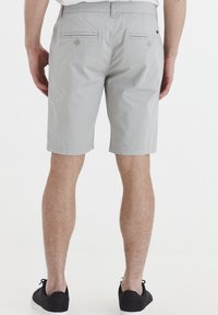 Shorts gris clair arrivant au genou en tissu lisse, dotés de deux poches arrière avec fermetures à bouton et d'un simple ourlet, associés à des chaussures noires.