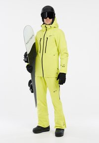 Veste et pantalon de ski jaune vif, gants noirs et casque. Tient une planche à neige avec une surface texturée et un logo.