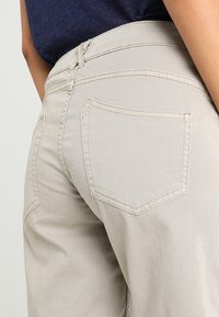 Beiga bomullshängselbyxor med hög midje, som har två bakfickor med böjda sömmar och en slät yta.