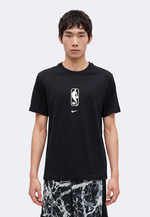 Ung mand iført en sort T-shirt med NBA-logo og sort-hvide mønstrede shorts, stående mod en ensfarvet lys baggrund.