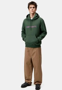 Haut à capuche vert avec logo violet, grande poche avant et cordon de serrage. Associé à un pantalon beige ample et des chaussures noires. Design simple et décontracté.