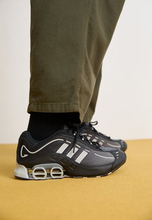 Black athletic shoes with white stripes, mesh upper, and visible cushioning. Paired with olive green pants, resting on a yellow surface.