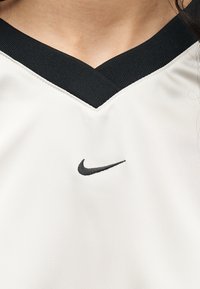 Άτομο που φορά αθλητική λευκή μπλούζα με V-λαιμόκοψη, μαύρο γιακά και μικρό μαύρο λογότυπο Nike Swoosh στο κέντρο κάτω από το λαιμόκοψη.