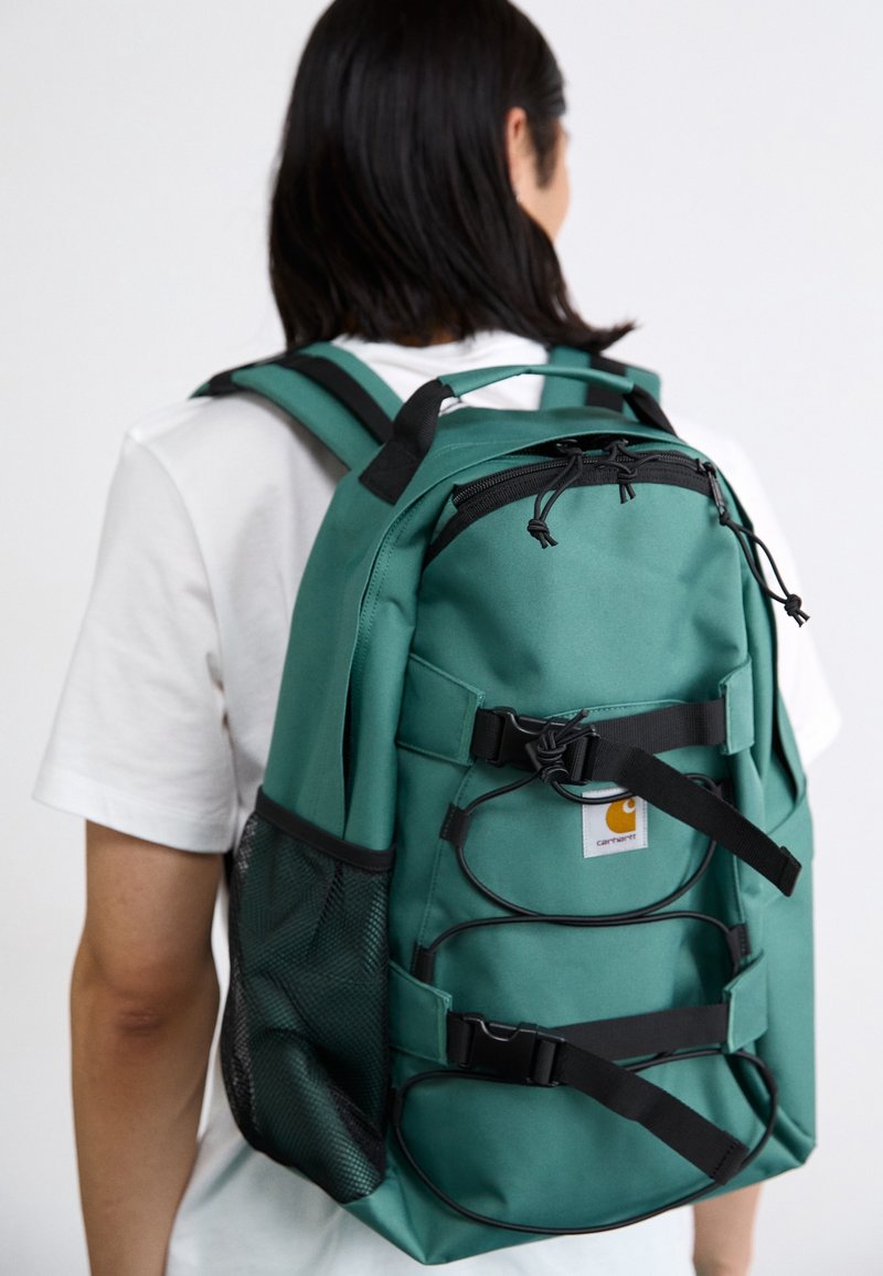 Persona con uno zaino verde con spallacci neri e tasca laterale in rete, mostrata da dietro su uno sfondo bianco semplice.