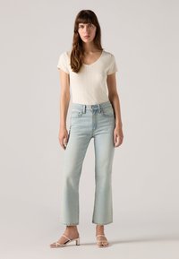 Jean flare bleu clair à taille haute, associé à un t-shirt crème à côtes et des sandales à talons blocs blanches à lanières. Textures lisses.