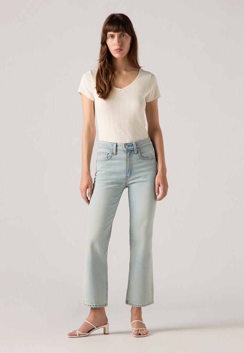 Jean flare bleu clair à taille haute, associé à un t-shirt crème à côtes et des sandales à talons blocs blanches à lanières. Textures lisses.