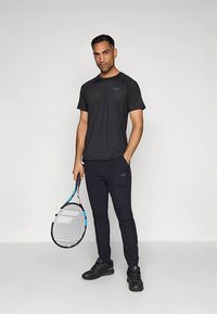 Man staat met een tennisracket in zijn rechterhand, draagt een zwart sportshirt, zwarte joggers en zwarte sneakers tegen een effen achtergrond.