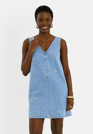 Mujer sonriente que lleva un vestido vaquero sin mangas de color azul claro con pequeño bordado floral, pendientes de aro dorados, anillos y una pulsera de oro.