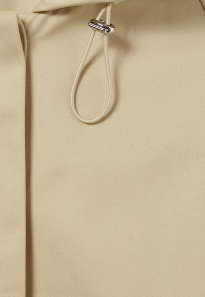 Tissu beige avec une texture lisse, présentant un cordon de serrage avec un embout métallique pour l'ajustement. Design simple et épuré avec des plis subtils.