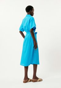 Blaues Kleid aus leichtem Stoff, mit Kragen, kurzen Ärmeln, einem Taillengürtel und einer mittellangen Silhouette. Trägt es mit braunen Sandalen.