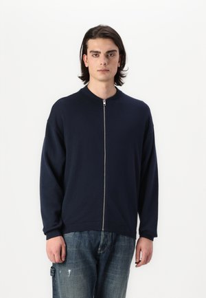 Marineblå glidelåsgenser med rund hals, avslappet passform, ribbestrikkede mansjetter og hem. Modellen har på seg slitte blå jeans.