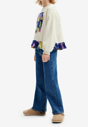 Sweatshirt beige clair avec un volan coloré au bas, associé à un jean bleu orné de petits points multicolores et des chaussures beiges.