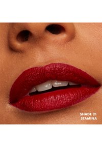 Rossetto opaco rosso applicato su labbra piene, che mostra una consistenza liscia. Ombra 21 "Stamina" etichettata nell'angolo in basso a destra.