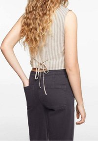 Béžový žebrovaný krátký top se zavazováním na zádech, v kombinaci s tmavě šedými kalhotami s dvěma zadními kapsami. Textura je hladká a přiléhavá.