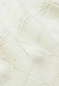 Texturovaná krémová látka s jemným vlnitým vzorem, měkkou texturou a viditelnými detaily šití. Materiál se jeví jako lehký a hladký.