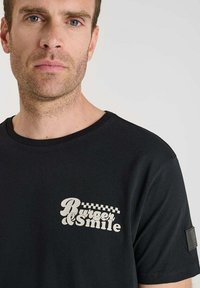 Camiseta de algodón negra con un gráfico en tonos crema que dice "Rubber Smile" con un detalle en cuadros en el lado izquierdo del pecho. Diseño de mangas cortas y cuello redondo.
