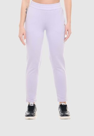 Persona che indossa pantaloni della tuta aderenti di colore lilla chiaro e sneakers nere, in piedi davanti a uno sfondo grigio chiaro semplice.