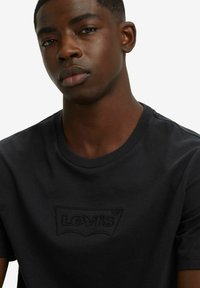 Camiseta de algodón negra con cuello redondo, que presenta un logo de Levi's en relieve en el pecho. Textura suave y diseño de ajuste regular.