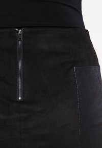 Jupe noire en matière suede texturée, avec une fermeture éclair à l'avant et un panneau en cuir contrastant sur le côté.