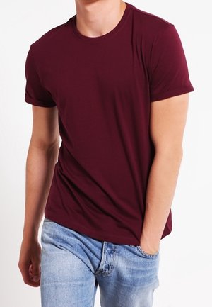 Homme portant un t-shirt bordeaux uni à manches courtes et un jean bleu clair, une main dans la poche, debout devant un fond blanc.