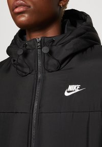 Veste rembourrée noire avec capuche, dotée d'une fermeture éclair à l'avant, de cordons de serrage et du logo Nike blanc sur la poitrine. Texture lisse, design décontracté.