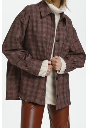 Persoon draagt een oversized bruine geruite jas over een crèmekleurige geribde coltrui en bruine leren broek, houdt de jas aan de voorkant gesloten.