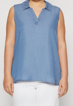 Top sin mangas de chambray azul claro con cuello en V y cuello, llevado con pantalones blancos en una persona contra un fondo liso.