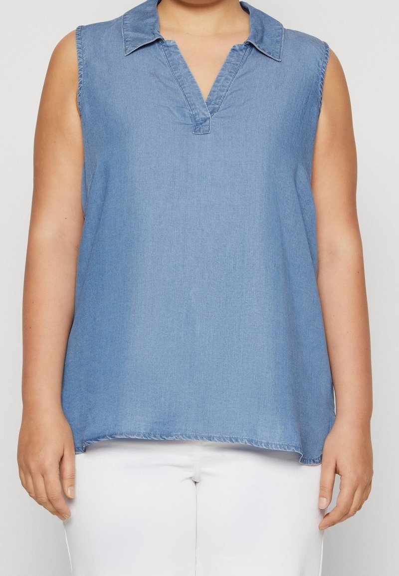Haut sans manches en chambray bleu clair avec col en V et col, porté avec un pantalon blanc sur une personne devant un fond uni.