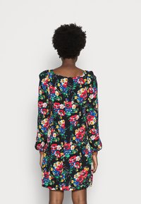 Vrouw met kort krullend haar die een zwarte jurk met kleurrijke rode, blauwe, gele en witte bloemenpatronen draagt, met de rug naar de kijker toegekeerd.