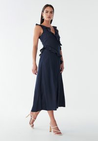 Navy blue maxi dress with ruffle details, a V-neckline, and a flowing design. Styled with white strappy heels on a neutral background.