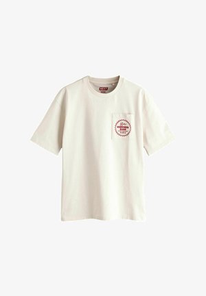 Camiseta color crema, de mangas cortas, con cuello redondo, con un estampado gráfico rojo en el bolsillo izquierdo. Fabricada en tejido de algodón.