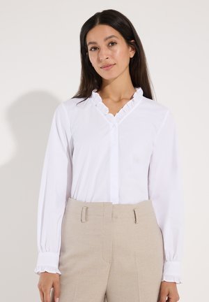 Vrouw draagt een witte blouse met lange mouwen en geruite kraag en manchetten, gecombineerd met beige broek, staand tegen een effen lichte achtergrond.